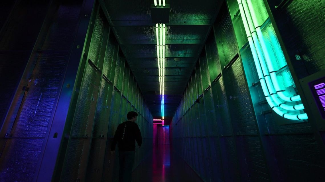 A guest walking a green-lit Portland hallway past a bundle of glowing tubes bending down its side, a pink doorway glowing at the far end.