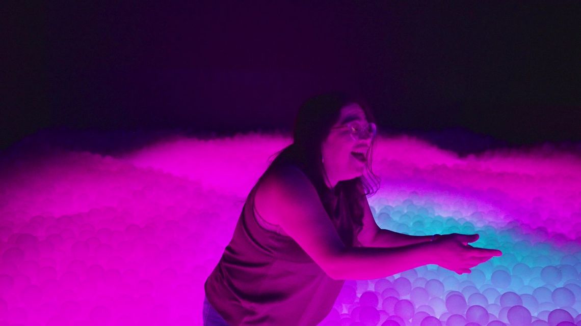 A guest laughing as she reaches into the pit, the balls glowing magenta around her and pale cyan under her hands.