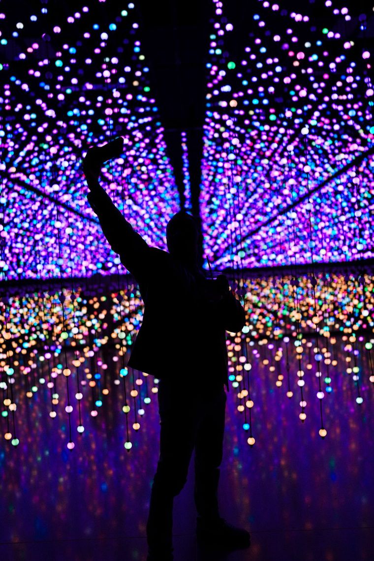 A guest in silhouette raises a phone overhead amid pink, blue and gold bulbs in Unknown Atmospheres.