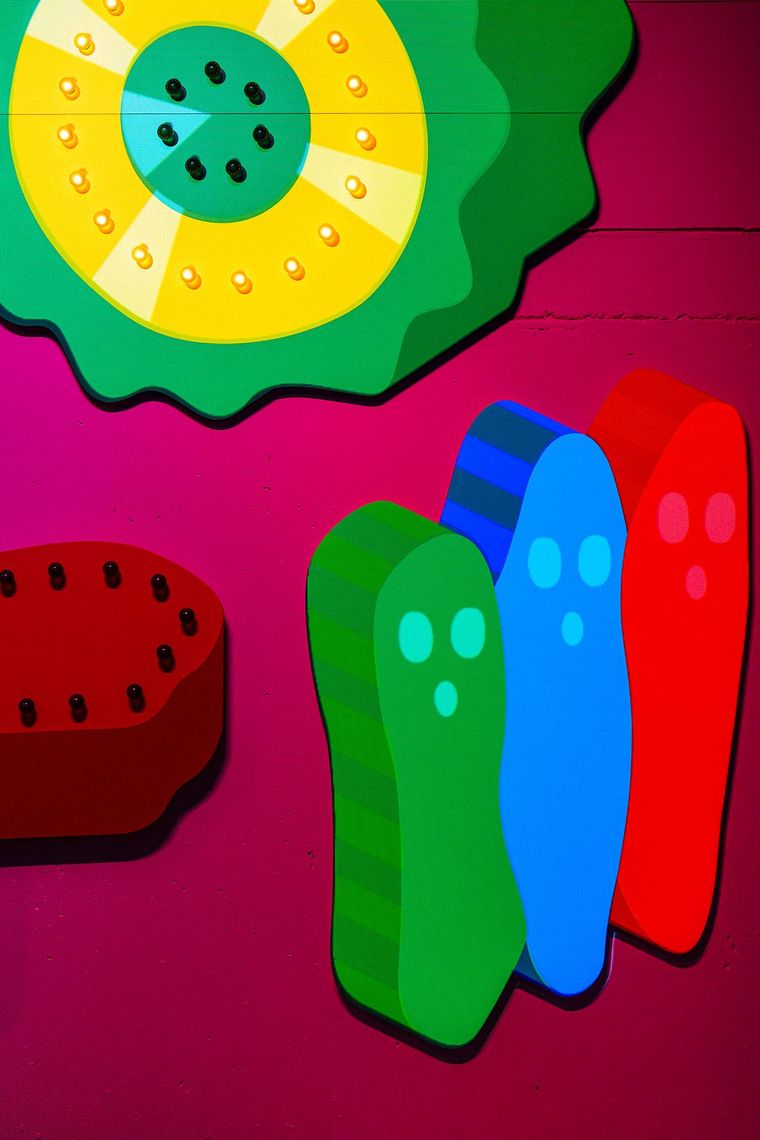 A yellow and green flower and three smiling bean shapes in green, blue and red on the pink wall of Are You There?