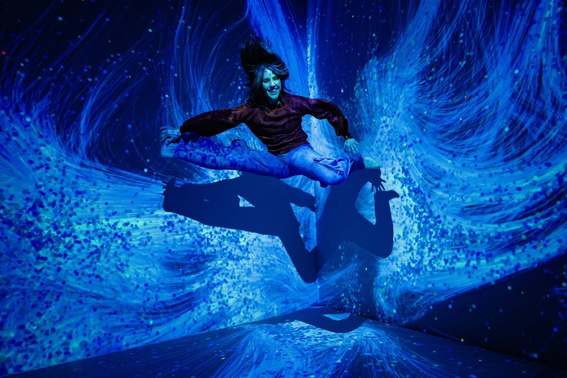 A guest leaps in midair, legs split, against swirling blue light in Quantum Trampoline.