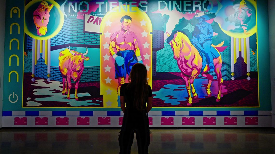 A guest standing before the No Tienes Dinero mural, a boxer, a bull and a rider on a horse in neon pinks, yellows and teal.