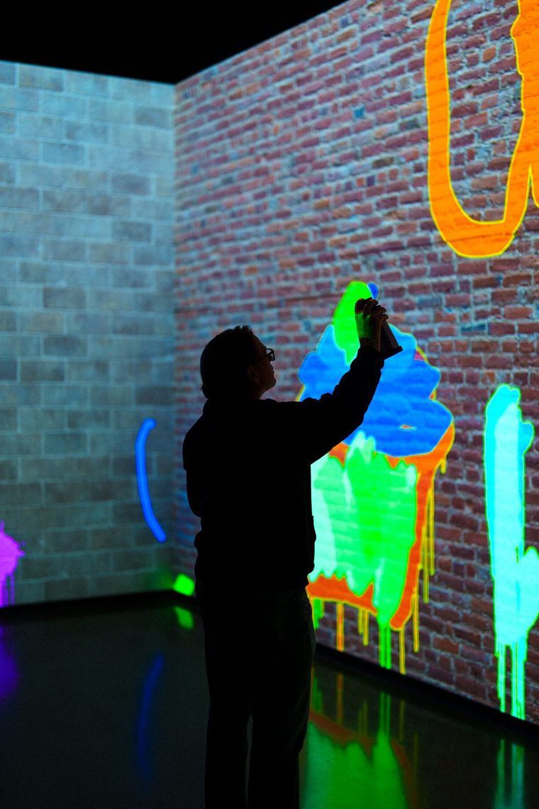 A guest raises a laser spray can in Laser Graffiti, drawing a dripping green and blue shape on projected brick.