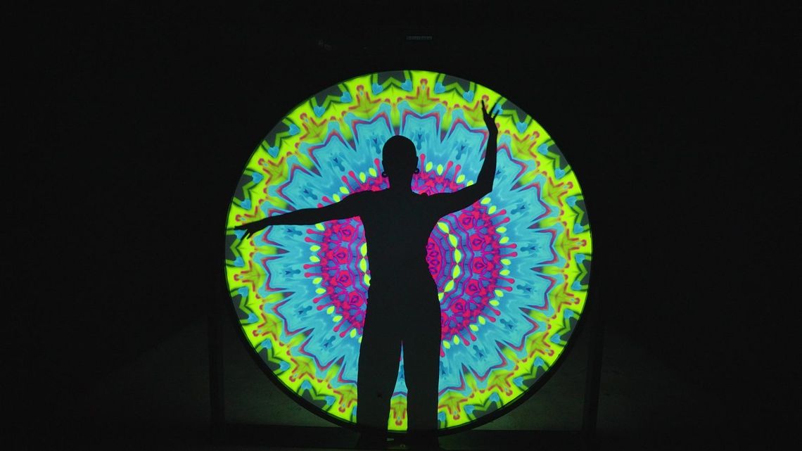 A guest in silhouette, one arm raised and one held out, against the disc of the Light Portal in lime, cyan and magenta.