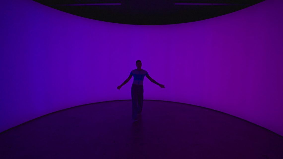 A small figure with arms out at the centre of the curved field of light, the whole chamber washed purple.