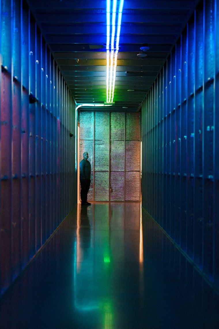 A blue hallway with a line of white light overhead and a backlit panel at its end, a guest small beside it.