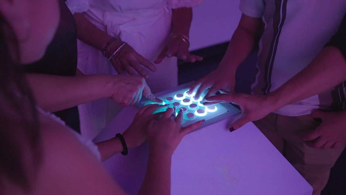 Several hands press the glowing button pad that sets the sculptures in motion.