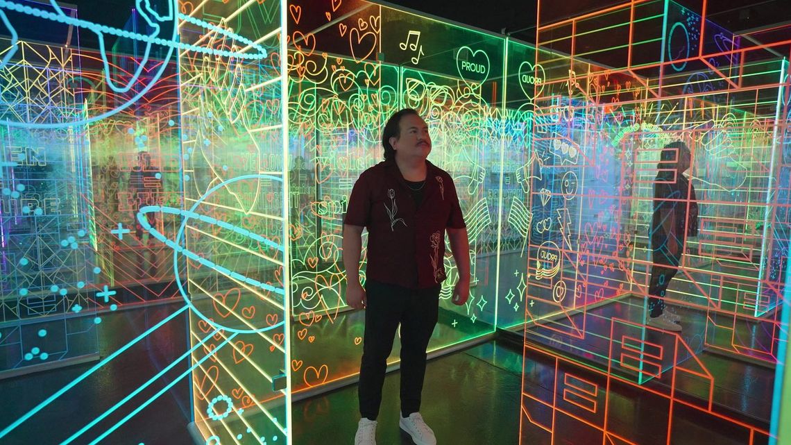 A guest standing among glowing acrylic panels in GAZE, San Antonio, looking up at etched hearts, planets and music notes in orange, green and blue.