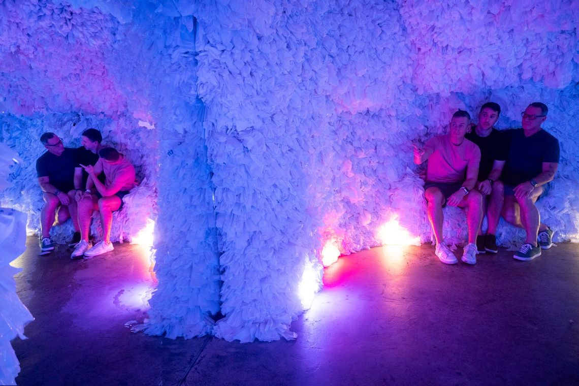 Guests sit on low ledges either side of a white column in Rainbow Cave, lit blue and pink.