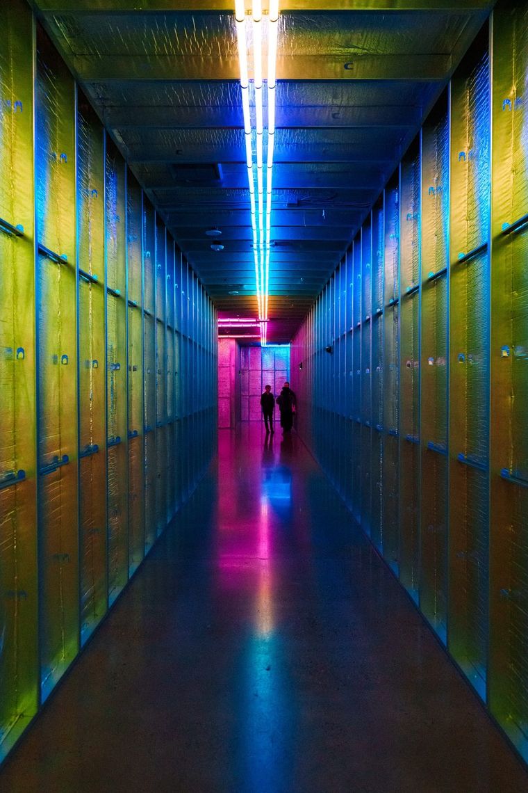 A long hallway in gold and blue light, a line of light down the ceiling, guests far down the corridor.