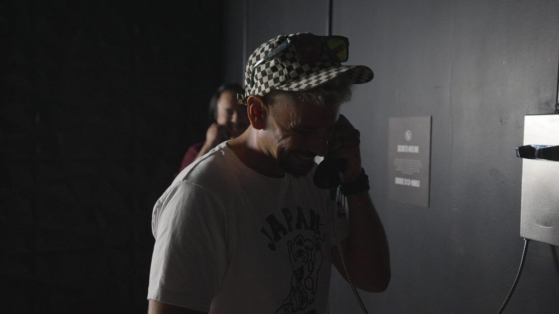 A guest in a checkered cap laughs into a phone, another guest behind him.