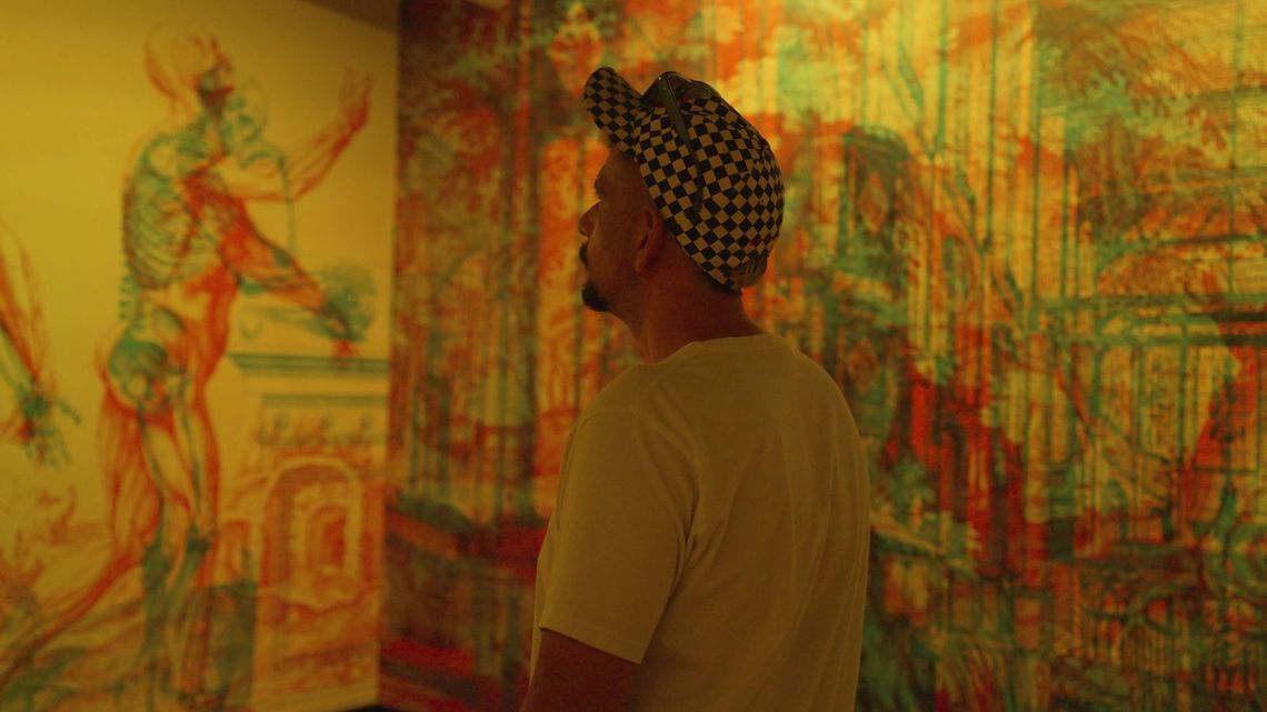 Under yellow light, an anatomical figure in the mural stands beside a guest in a checkered cap.