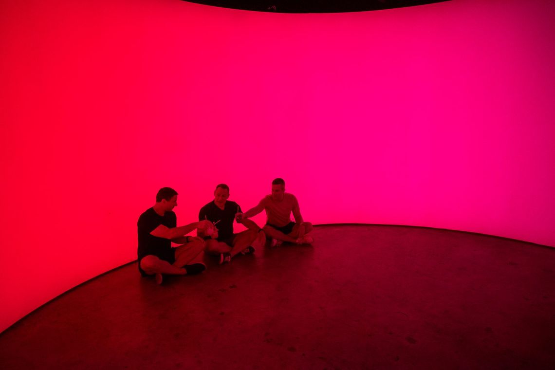 Three guests sit cross-legged on the floor and toast with their drinks as the light glows red into pink.