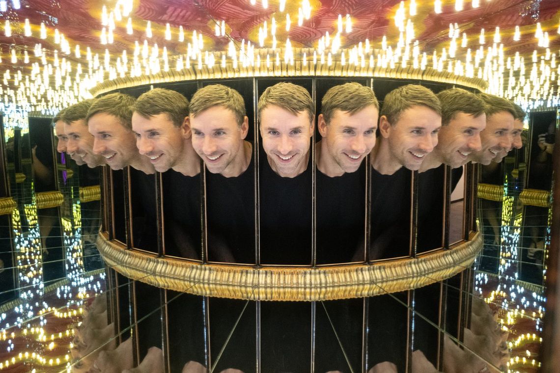 A smiling guest repeated in a ring of mirrors inside Infinity Box, a gold rail below and rows of bulbs above.
