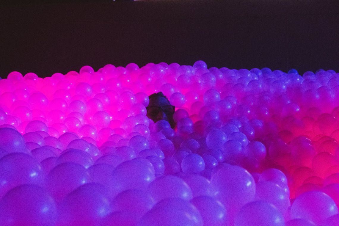 A guest's face peeks out from deep in the Diodic Daydream ball pit, the balls lit pink and violet.