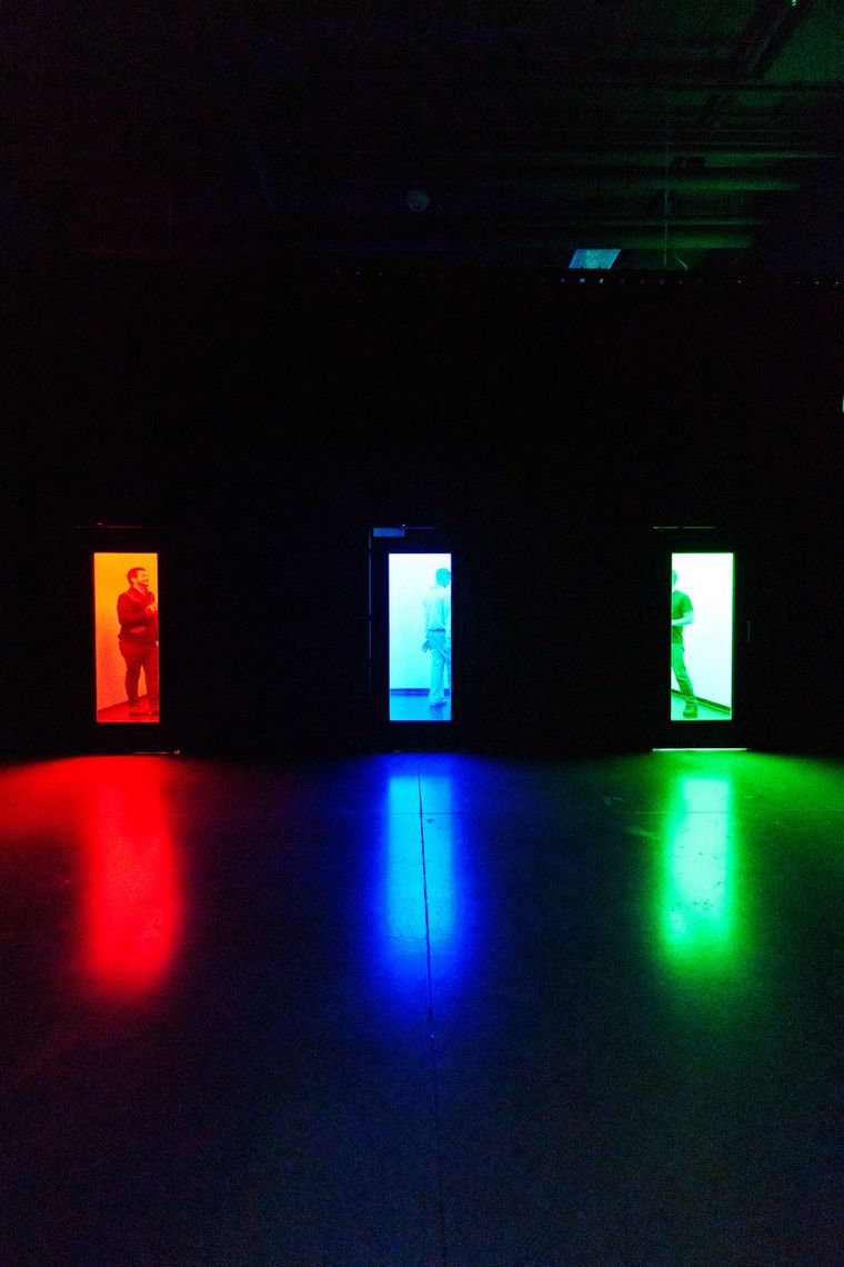 Three lit doorways in Chromesthesia, red, blue and green, a guest in each, their colors reflected on the floor.