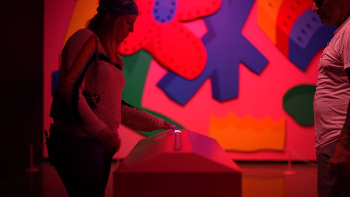 A guest presses a button on the console in Are You There?, bright painted shapes on the wall behind her.