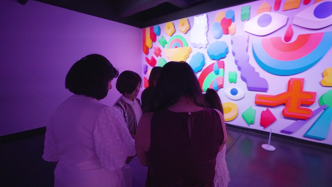 A group of guests seen from behind before the lit face of Happiness, its eyes, rainbows and cut-out shapes in bright colour.