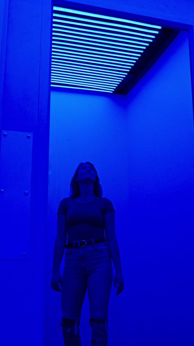 Inside the blue chamber of Chromesthesia, a guest stands in a wash of blue light, looking up toward the glowing slats overhead.