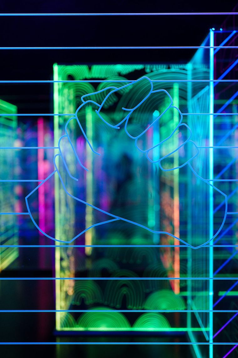 Two clasped hands etched in blue on an acrylic panel in GAZE, green and pink panels glowing behind.