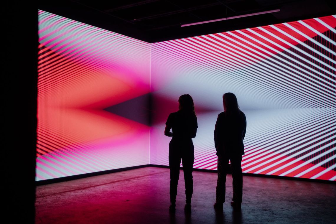 Two guests in silhouette face a corner where red and white stripes of light meet in Onion Skin.