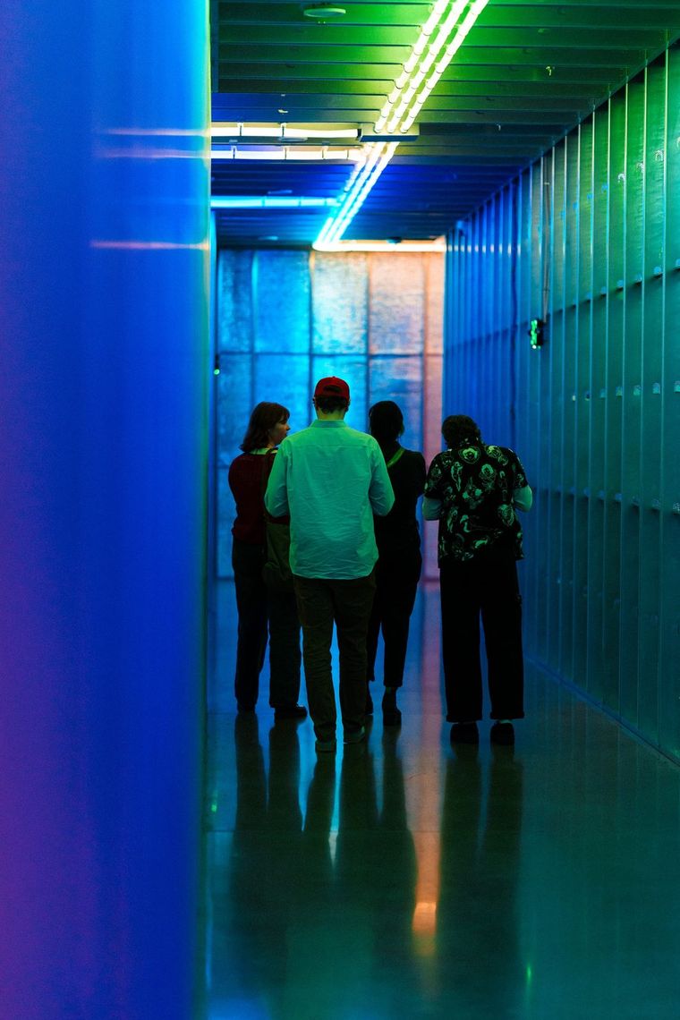 Three guests in a teal hallway under a rainbow line of light, a lit panel ahead of them.