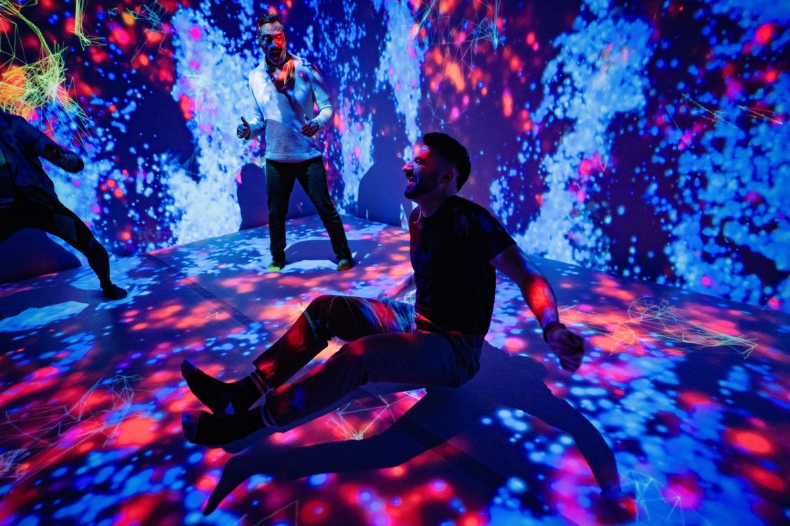 Two guests play in Quantum Trampoline, one sitting back on a floor scattered with pink and blue light.