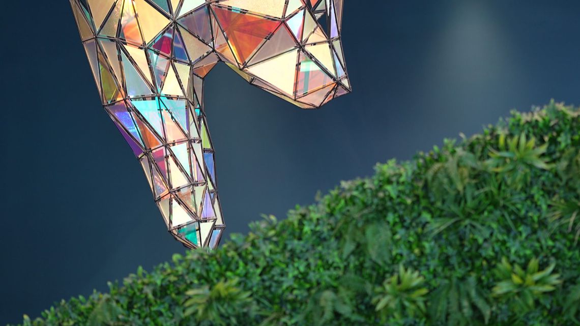 The iridescent faceted sculpture leaning down until its muzzle meets the green hedge, the panels catching orange, violet and teal.