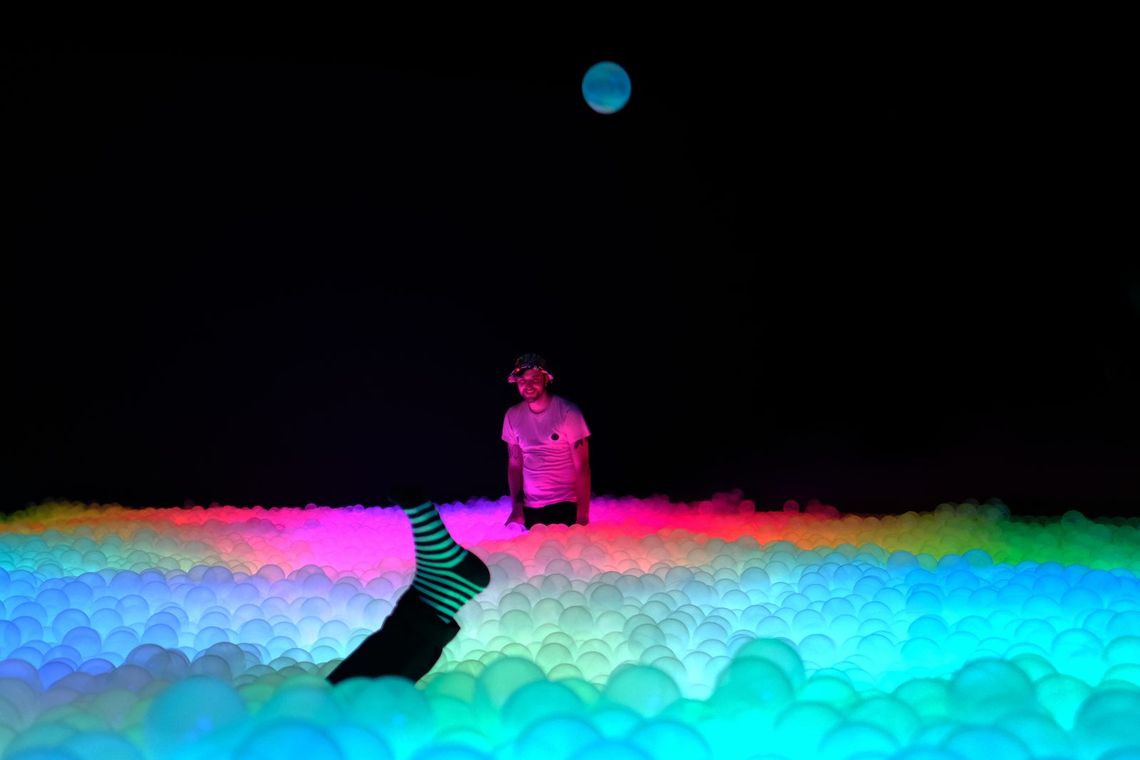 A guest dives into the ball pit, legs in striped socks in the air, the balls lit cyan, pink and green.