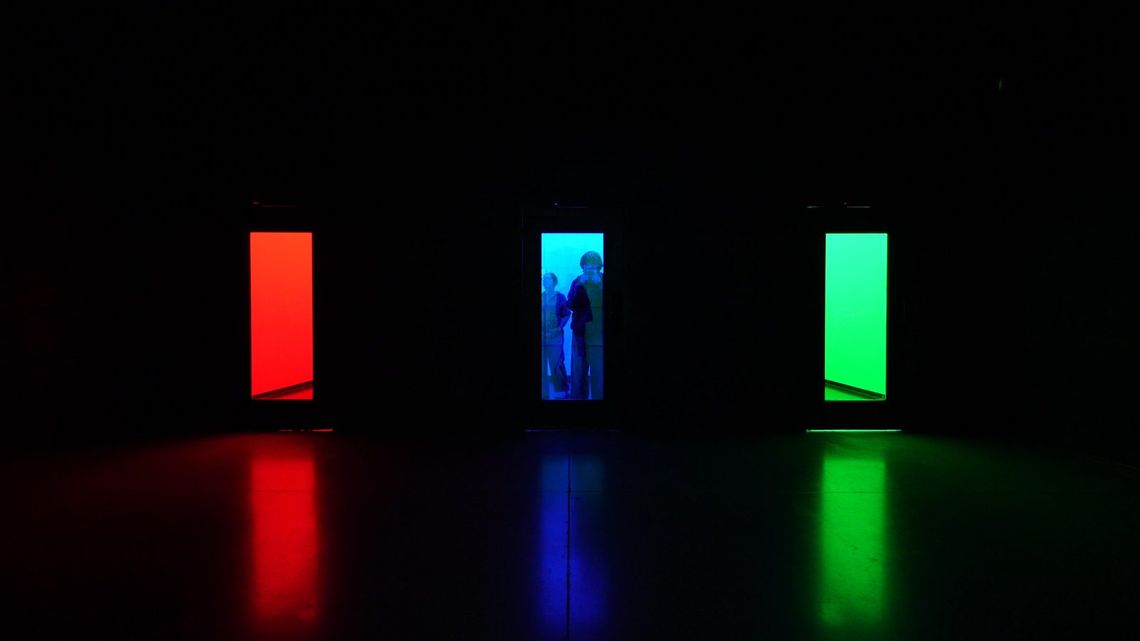 Three lit doorways in the dark, red, blue and green, their colours pooling across the floor, one small silhouette in the blue door.