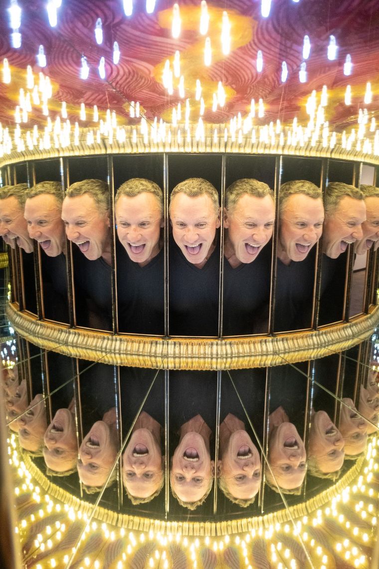 Inside Infinity Box a laughing guest repeats around a mirrored ring under a ceiling of chandelier lights.
