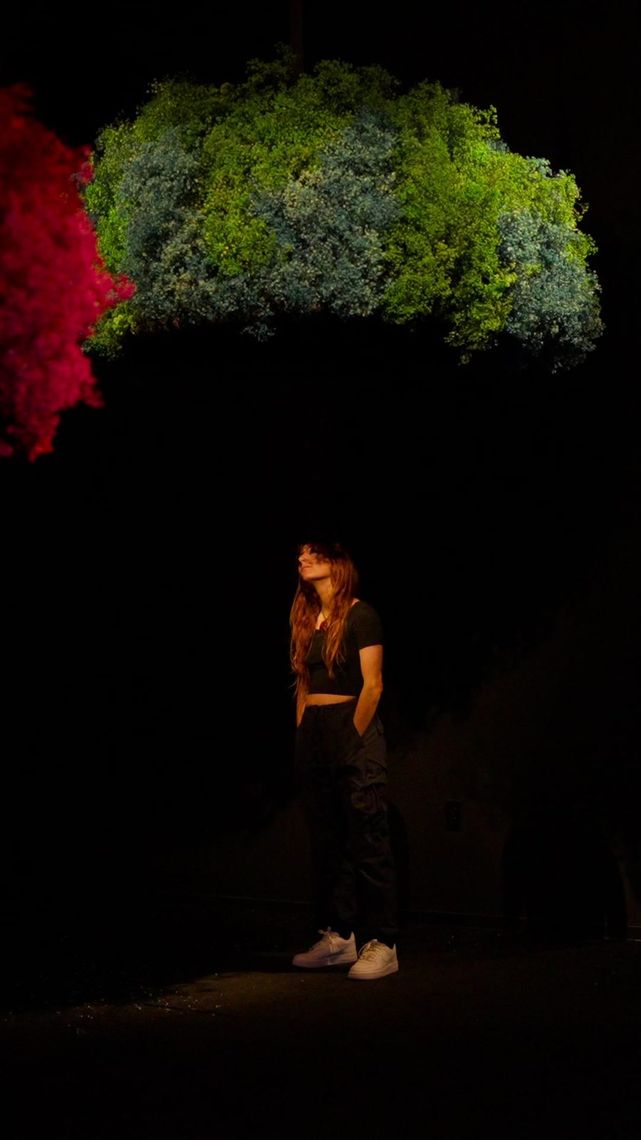 A guest standing in a pool of light beneath a hanging canopy of blue-green flowers, a red canopy at the edge of the frame.