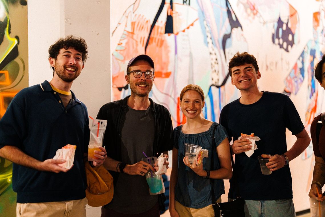 Four people holding drinks and smiling in front of a painted gallery wall at a partner night
